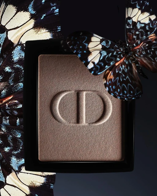Close-up of Dior eyeshadow palette with CD logo, adorned with butterfly. Studio shot showcases beauty product with natural elements.