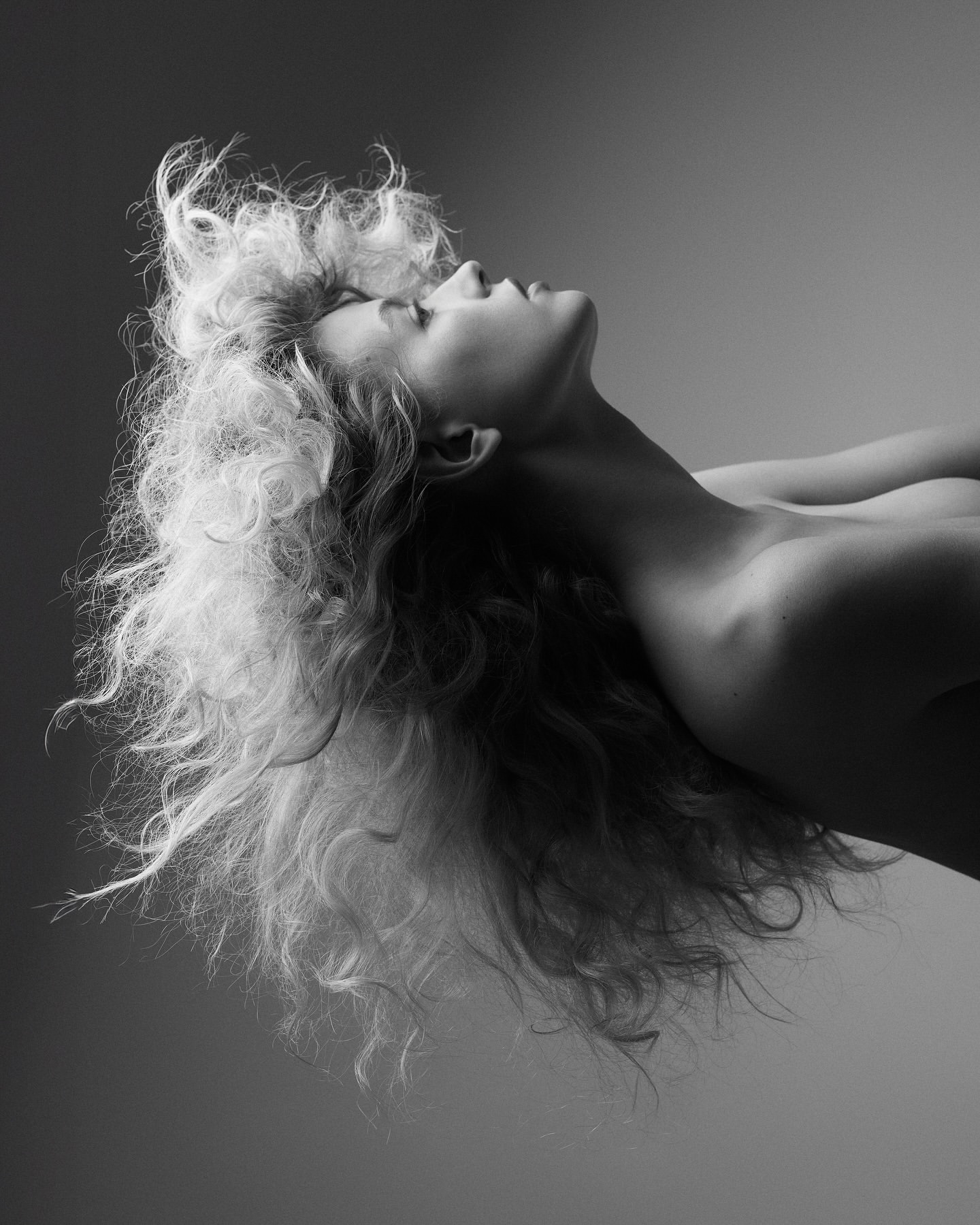 Black and white portrait of a fair-skinned adult woman with curly blonde hair, looking up with her head tilted back in a studio setting.