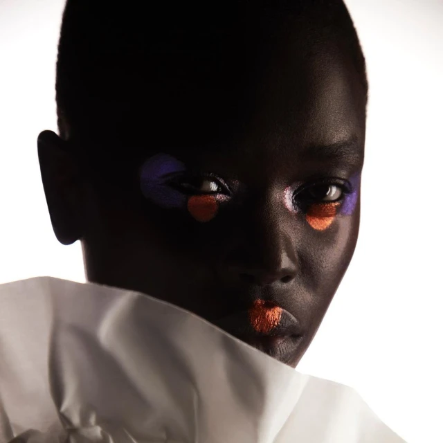 Close-up portrait of a black woman with dark hair and bold makeup with orange lips and a purple and orange circle under her eyes.