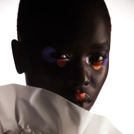 Close-up portrait of a black woman with dark hair and bold makeup with orange lips and a purple and orange circle under her eyes.