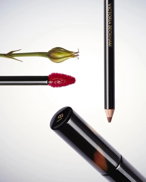 Victoria Beckham Beauty collection featuring lipstick, lip liner, eyeliner, and a green bud on a white background for cosmetics advertising.