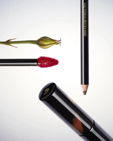 Victoria Beckham Beauty collection featuring lipstick, lip liner, eyeliner, and a green bud on a white background for cosmetics advertising.
