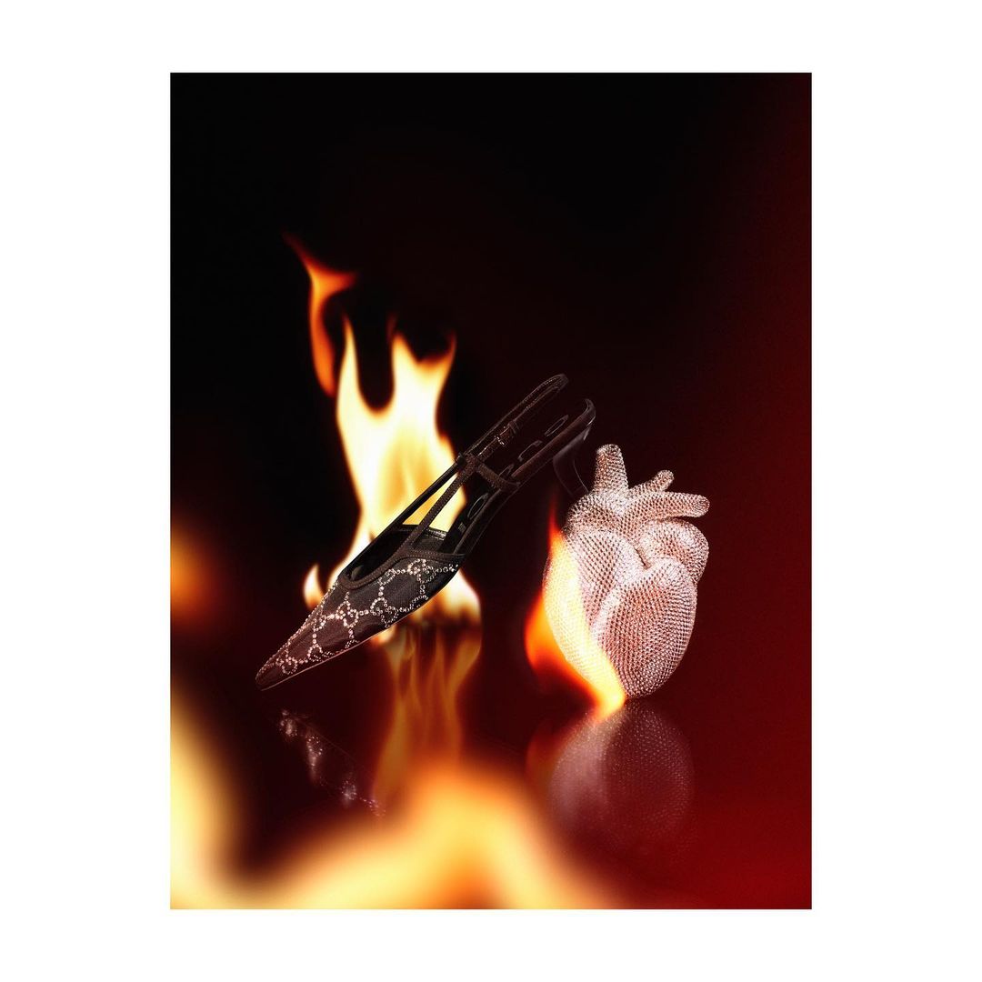 High-heeled shoe with logo and a decorative heart next to flames against a black background in a dramatically lit studio shot.