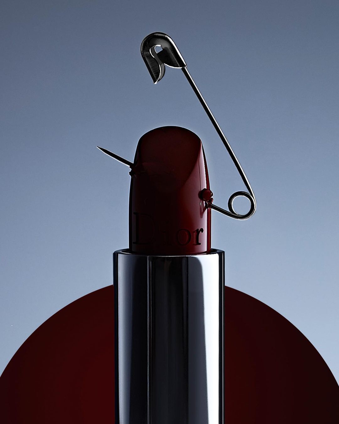 Still life close-up of a red lipstick tube pierced with a safety pin against a soft blue background in studio lighting.