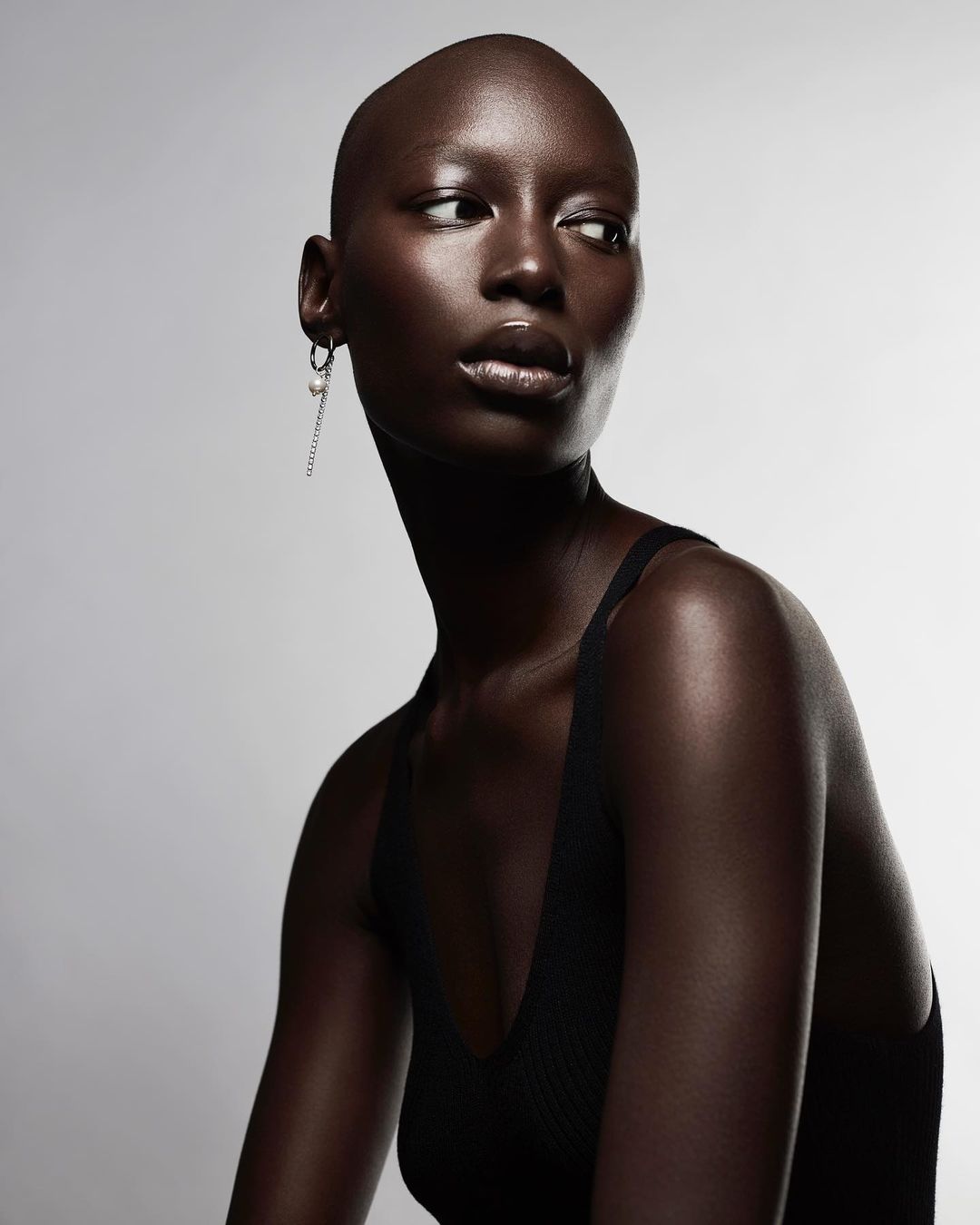 Striking portrait of a bald, dark-skinned woman with an earring, wearing a black tank top against a simple, pale gray backdrop.