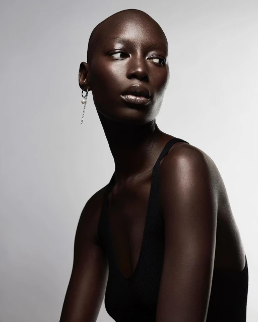 Striking portrait of a bald, dark-skinned woman with an earring, wearing a black tank top against a simple, pale gray backdrop.