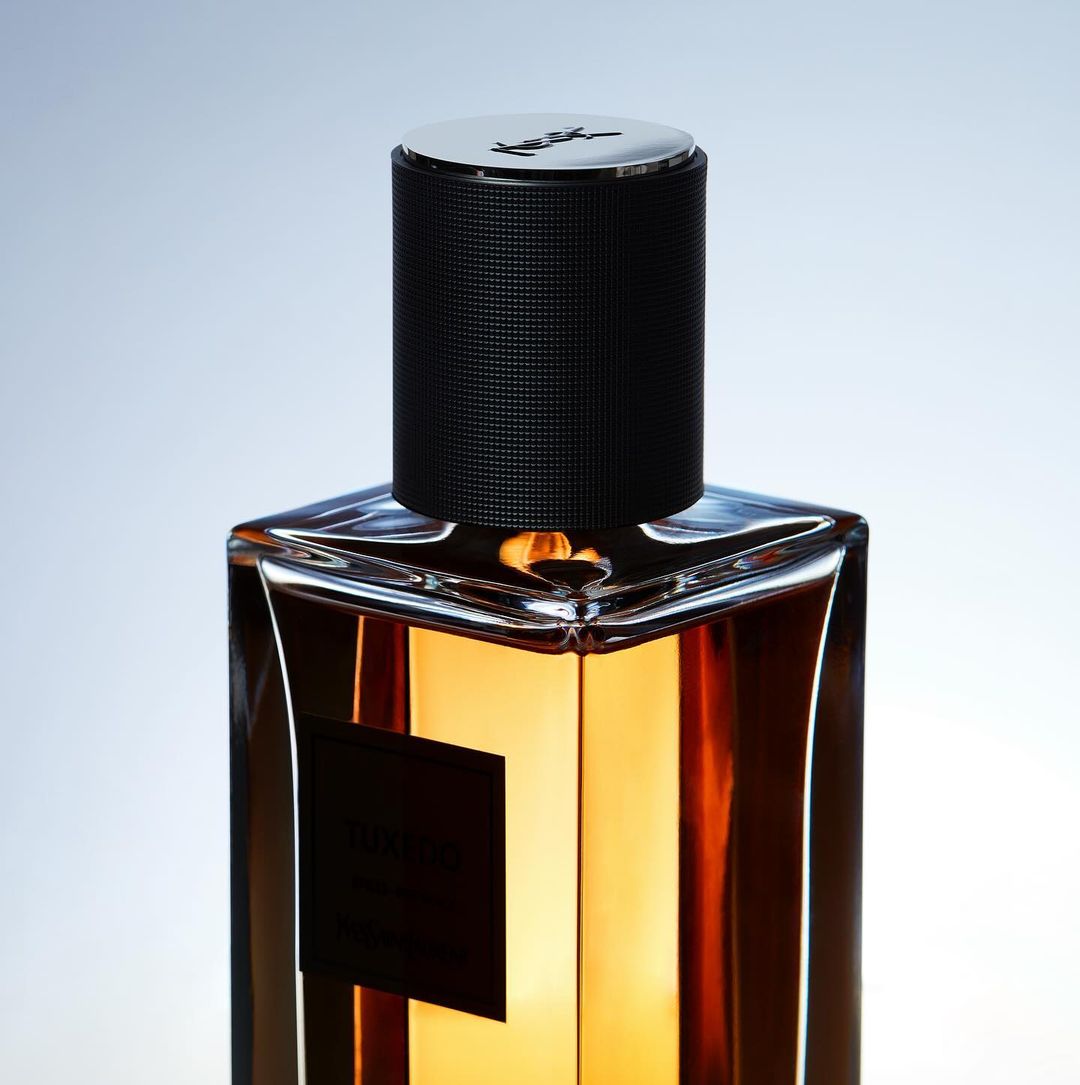 Product shot of a glass perfume bottle with brown liquid, black textured cap and silver top, against a soft blue and white gradient background.