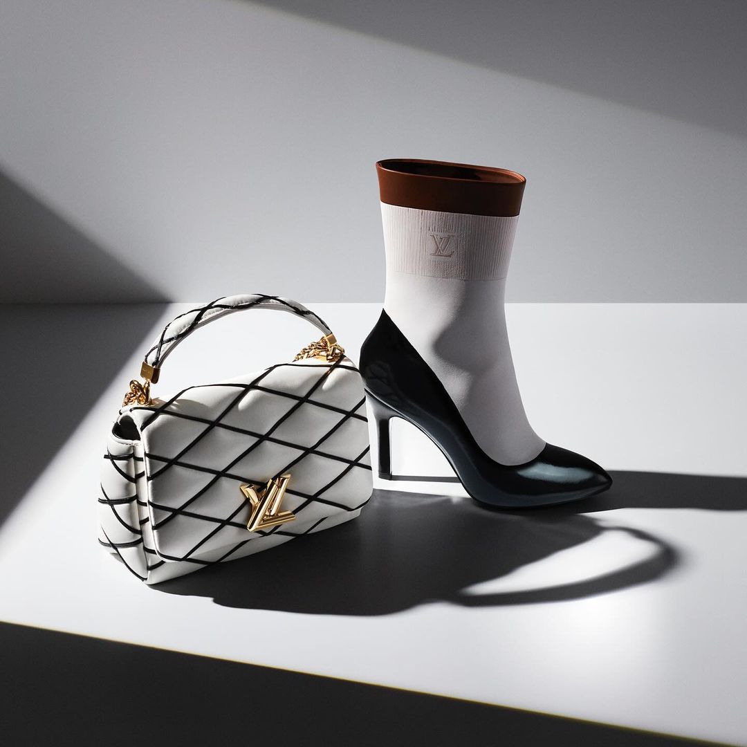 High heel shoe, white quilted purse with gold logo, fashion product photography in a brightly lit studio setting.