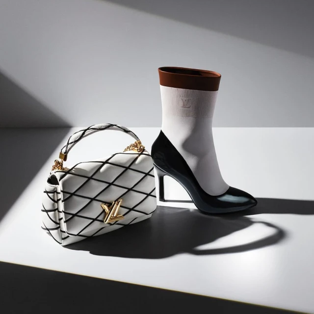High heel shoe, white quilted purse with gold logo, fashion product photography in a brightly lit studio setting.