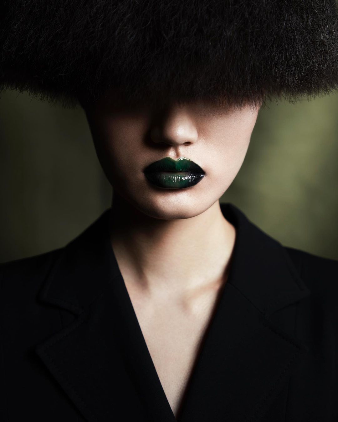 Asian female fashion portrait with oversized dark afro, wearing a black jacket with strong green glossy lipstick against a blurred background.