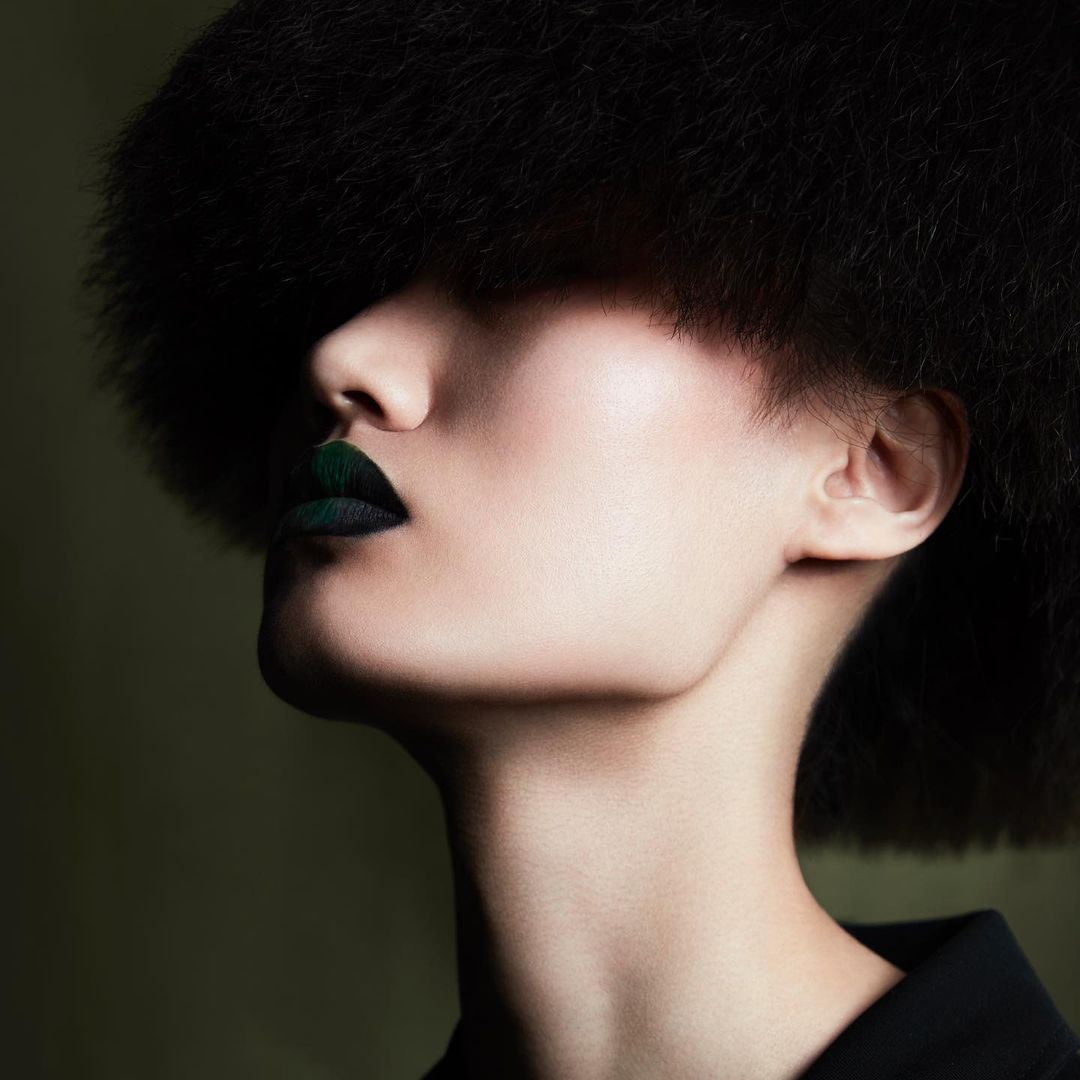 Woman with dark hair and dramatic dark green lipstick, eyes shadowed, in a close-up studio beauty shot with a dark background.