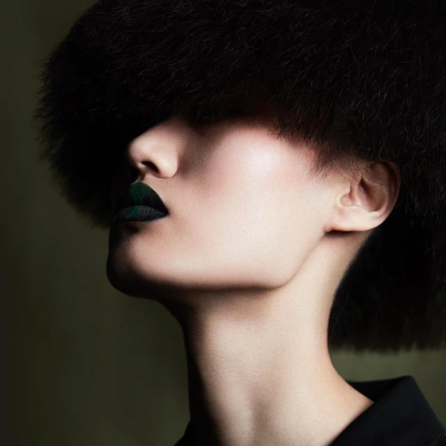 Woman with dark hair and dramatic dark green lipstick, eyes shadowed, in a close-up studio beauty shot with a dark background.