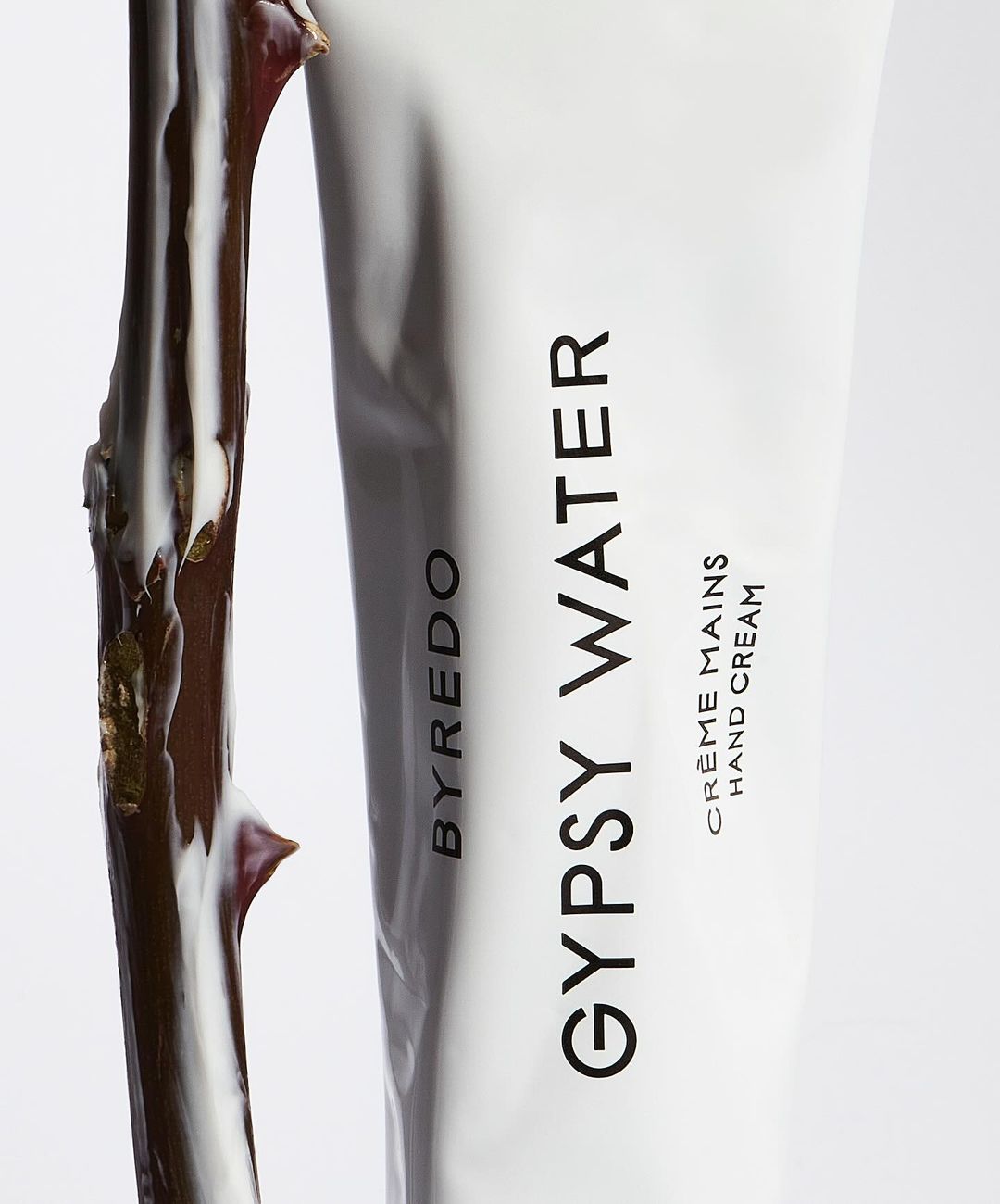 Byredo Gypsy Water hand cream tube next to a branch. Close-up studio shot with white background. Beauty and wellness product.