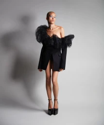 Model with shaved blonde hair in a black blazer with sheer tulle ruffles standing against a grey studio backdrop