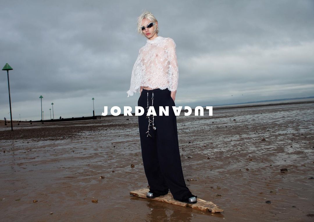 Model on a wet beach wearing a white lace top, black wide-leg pants, and sunglasses stands on a piece of driftwood in an editorial fashion shot.
