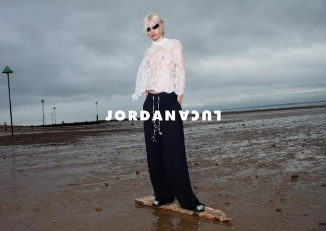 Model on a wet beach wearing a white lace top, black wide-leg pants, and sunglasses stands on a piece of driftwood in an editorial fashion shot.