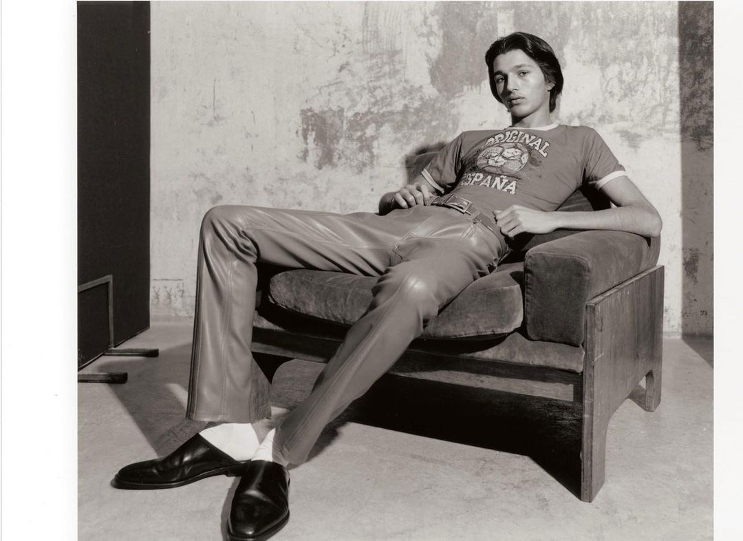 Indoor portrait of a reclining adult male wearing leather pants, t-shirt and black shoes sitting in a velvet chair, and plaster wall background.