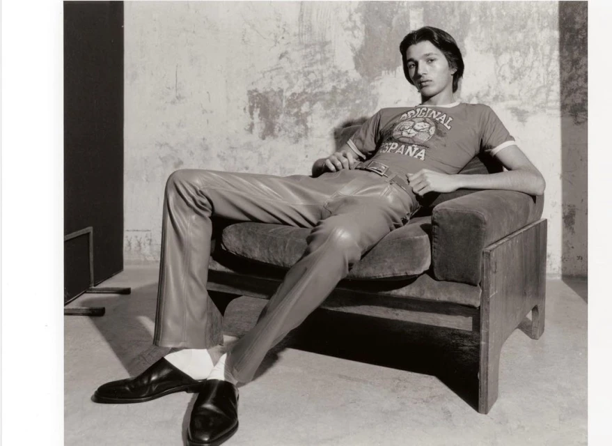 Indoor portrait of a reclining adult male wearing leather pants, t-shirt and black shoes sitting in a velvet chair, and plaster wall background.