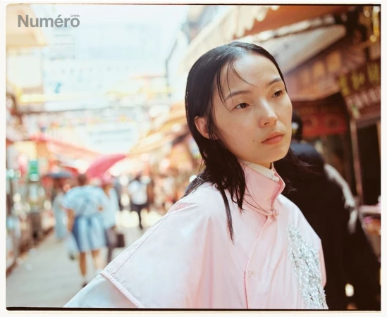 Editorial fashion shot of a young Asian woman in a pink jacket, walking along a crowded city street with shopfronts, in natural light.