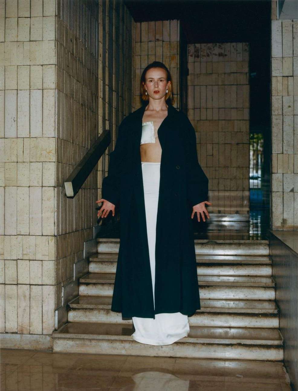 A young woman stands on stone steps in a long black coat and white outfit in an urban environment with neutral colors.