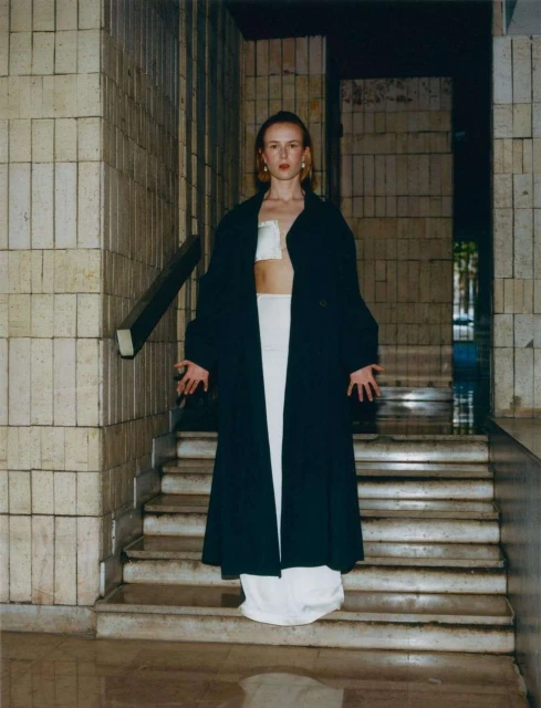 A young woman stands on stone steps in a long black coat and white outfit in an urban environment with neutral colors.