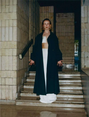 A young woman stands on stone steps in a long black coat and white outfit in an urban environment with neutral colors.