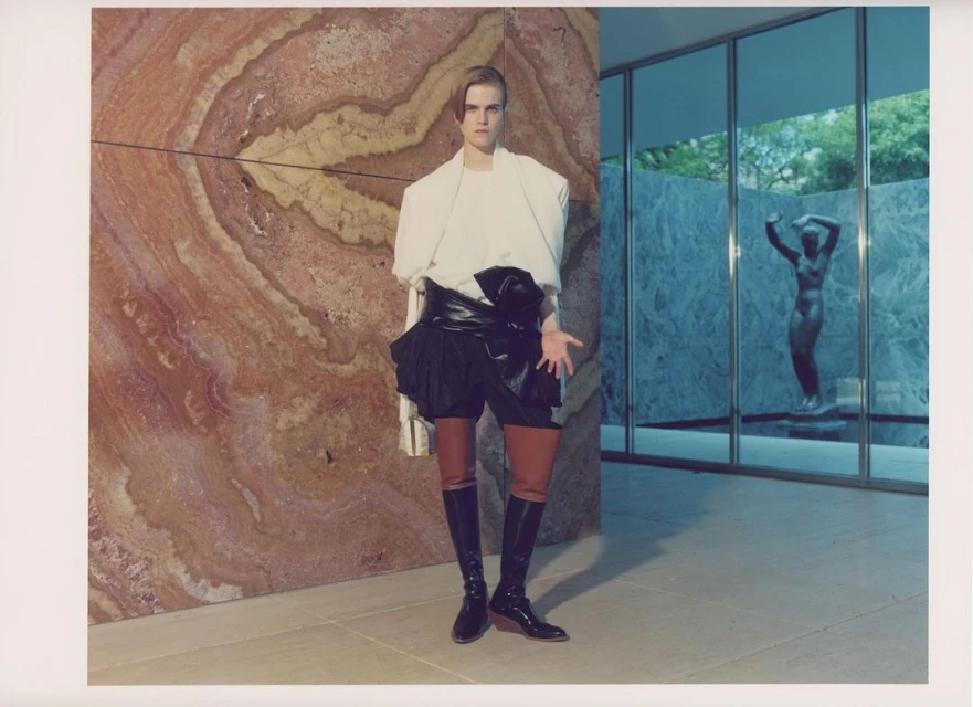 Fashion shot featuring a person in a white top, black shorts, and long black boots, posing indoors against a textured marble wall.