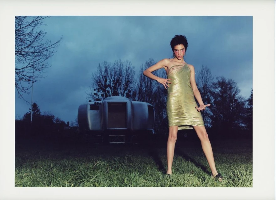 An adult female fashion model poses in a gold dress outdoors against a dark blue sky with a futuristic RV-style camper in the background.