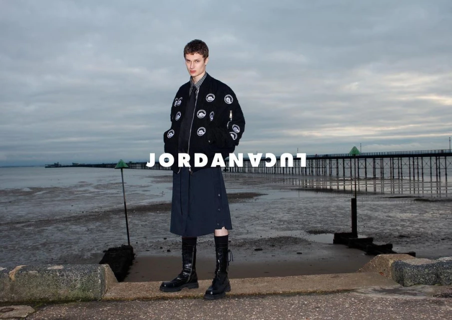 Male model in a black bomber jacket and skirt stands on a beach with a pier in the background, styled for a fashion editorial.