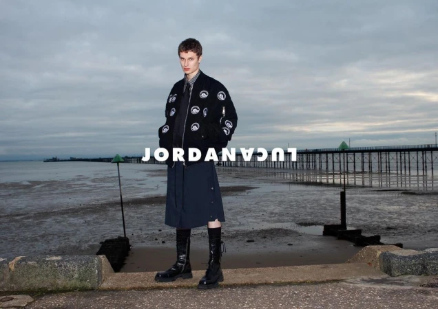 Male model in a black bomber jacket and skirt stands on a beach with a pier in the background, styled for a fashion editorial.