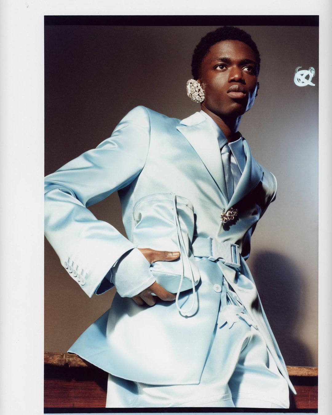 Portrait of a young Black male fashion model in a light-blue suit with a tie and jewelry, posing indoors in front of a grey wall.
