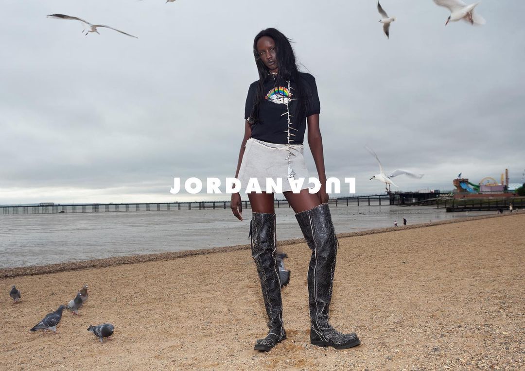 Fashionable adult female poses on a sandy beach with gulls in flight, wearing a black top, mini skirt, and thigh-high boots under an overcast sky.