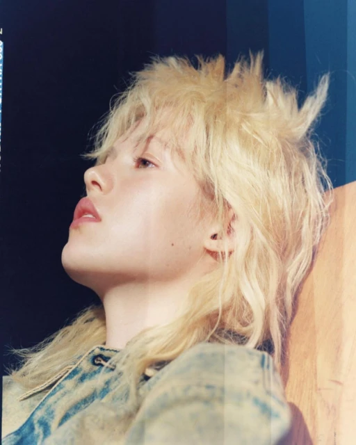 Close-up side profile of a blonde woman with spiky hair, wearing a denim jacket against a dark background with blue and orange accents.