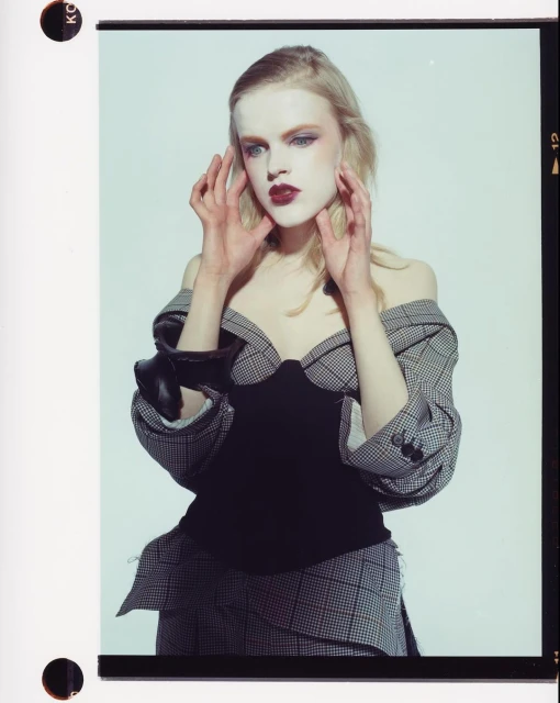 Blonde model in a black bustier and off-the-shoulder plaid jacket strikes a pose in a studio with her hands near her face.