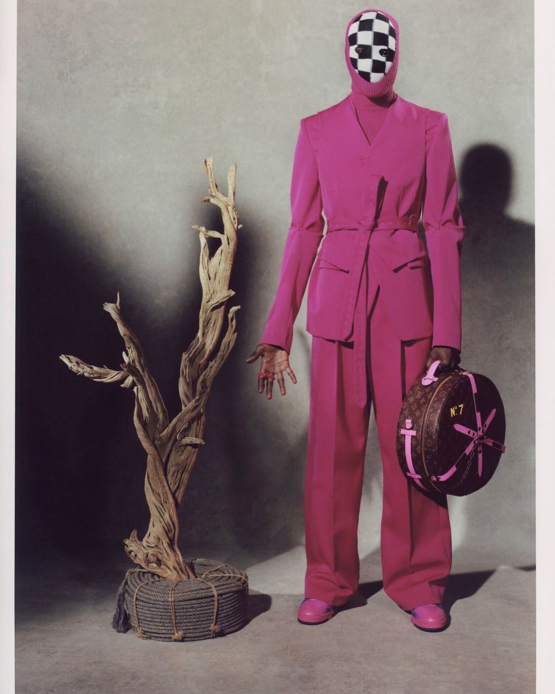 Model in pink suit and checkerboard balaclava poses with tree and designer bag, creating a surreal fashion editorial image.