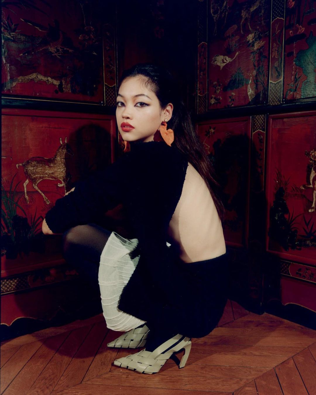 Asian woman in a black outfit poses in a squat position, wearing woven heels against an ornate red panelled wall and wood floor.