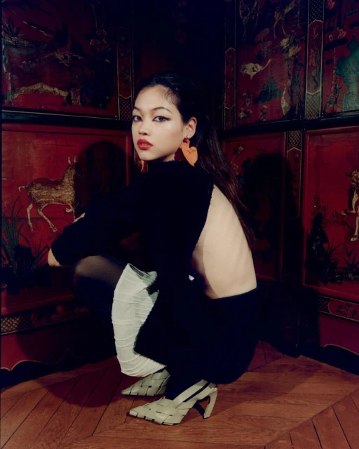 Asian woman in a black outfit poses in a squat position, wearing woven heels against an ornate red panelled wall and wood floor.