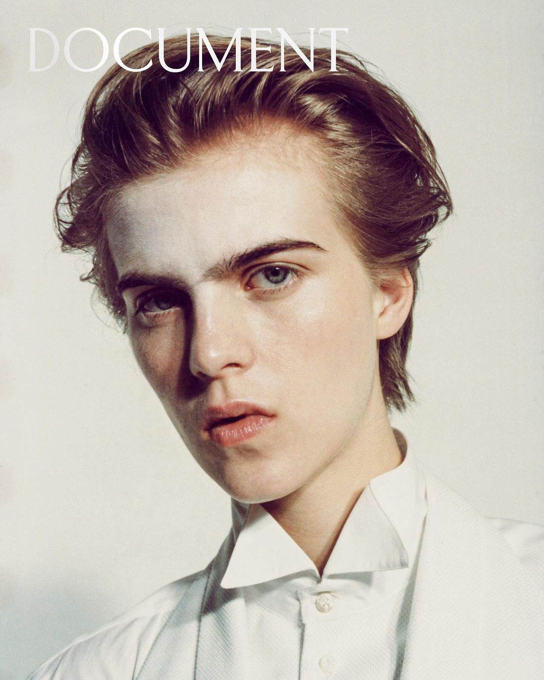Close-up portrait of a young adult male with light skin and light brown hair, wearing a white button-down shirt and white make-up on the face.