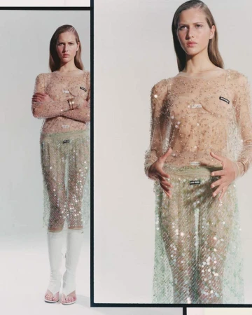 Fashion shot of a slender woman in a sheer, embellished long-sleeved dress with white boots, posing against a neutral studio background with reflection.