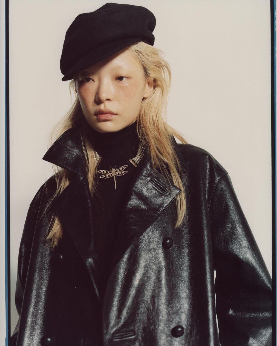 Portrait of a young woman with fair hair wearing a black newsboy cap, a black turtleneck, a dragonfly pendant necklace, and a black leather coat.
