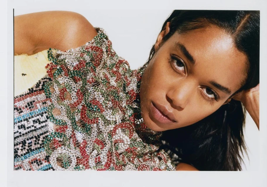 Portrait of a model with dark hair posing with her hand behind her head wearing a beaded high neck garment in a white photo studio.