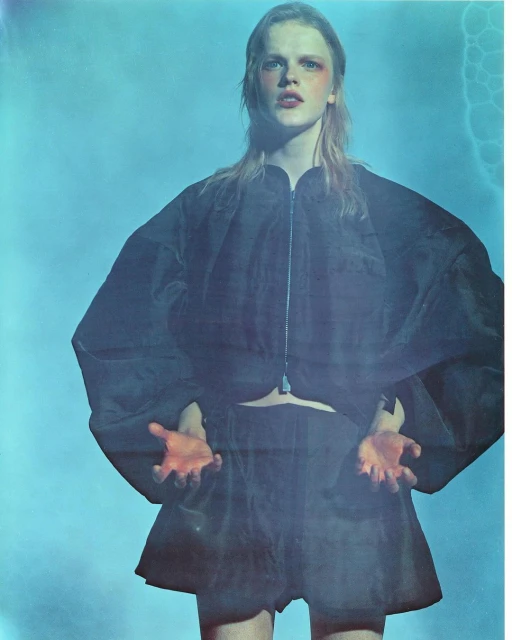 Young woman in black avant-garde fashion stands with outstretched hands against a hazy blue background with digital lightning effects.