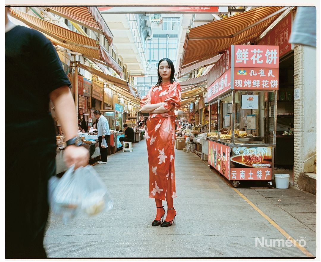 Fashionable young woman poses in a patterned dress and high heels in a busy Asian marketplace, surrounded by stalls and people.