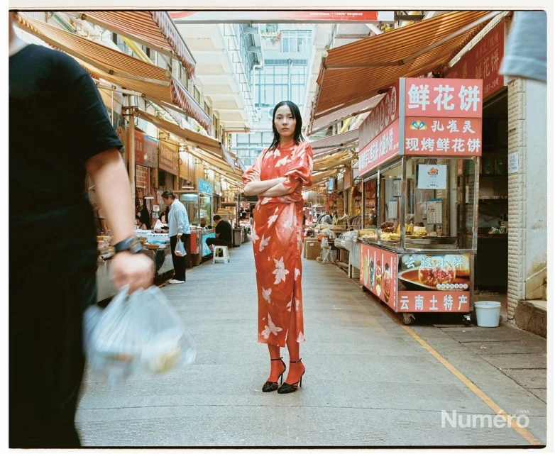 Fashionable young woman poses in a patterned dress and high heels in a busy Asian marketplace, surrounded by stalls and people.