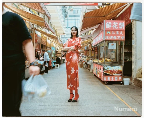 Fashionable young woman poses in a patterned dress and high heels in a busy Asian marketplace, surrounded by stalls and people.
