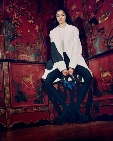 Model wearing a white and black coat, dark tights, and platform heels, posing against ornate red panel walls.