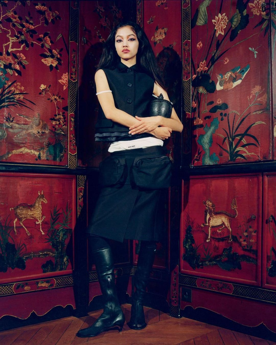 A full portrait of a young woman in a black vest and skirt holding a basket, posing in front of a red oriental screen in a studio.
