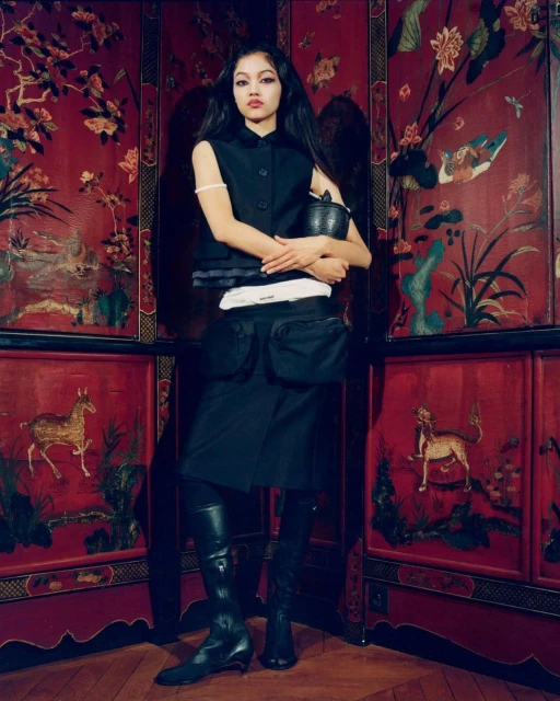 A full portrait of a young woman in a black vest and skirt holding a basket, posing in front of a red oriental screen in a studio.