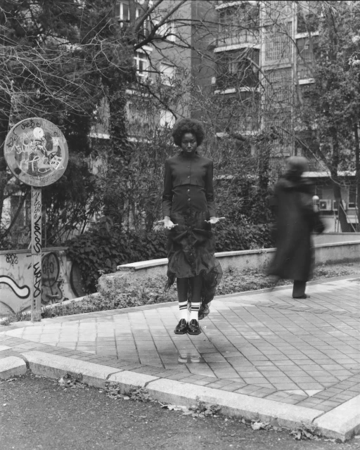 Black and white shot of a Black woman suspended in the air, mid-jump, wearing a dark dress and striped socks with a street scene background.