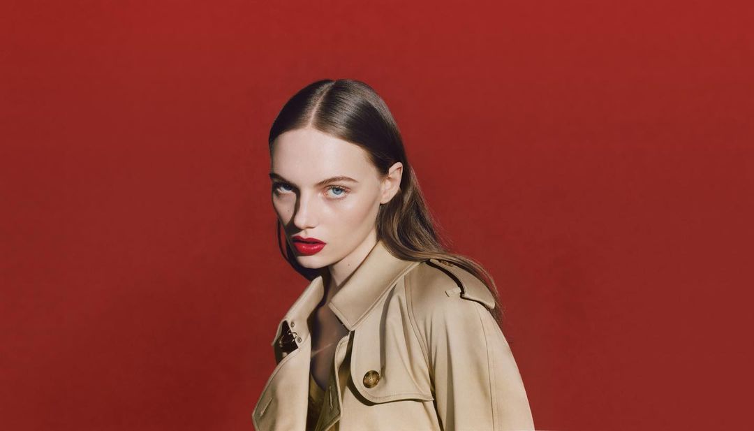 Portrait of a young adult woman with neutral expression in tan coat against red backdrop. Fashion, beauty and portrait photography.